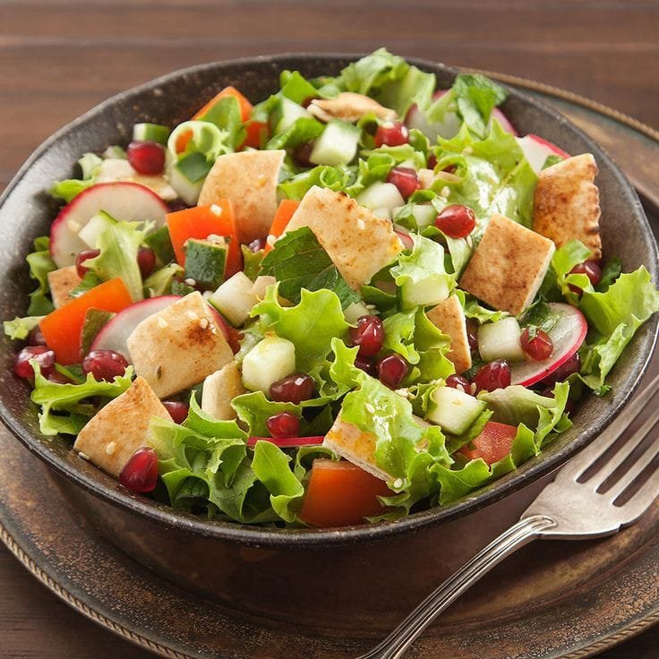 Ensalada fresca y colorida en un plato oscuro, compuesta de lechuga, pepino, rábanos, zanahorias, pan tostado en cubos y granos de granada, acompañada de un aderezo ligero. Una de las Recetas Saludables para Diabéticos