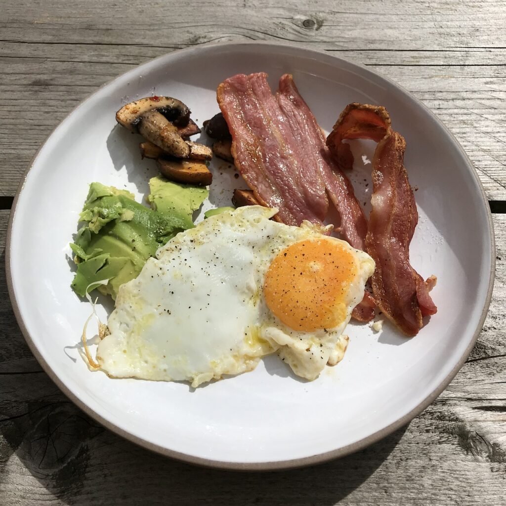 Desayuno de Dieta Cetogénica con huevo frito, aguacate, tocino crujiente y champiñones salteados en un plato blanco sobre una mesa rústica de madera